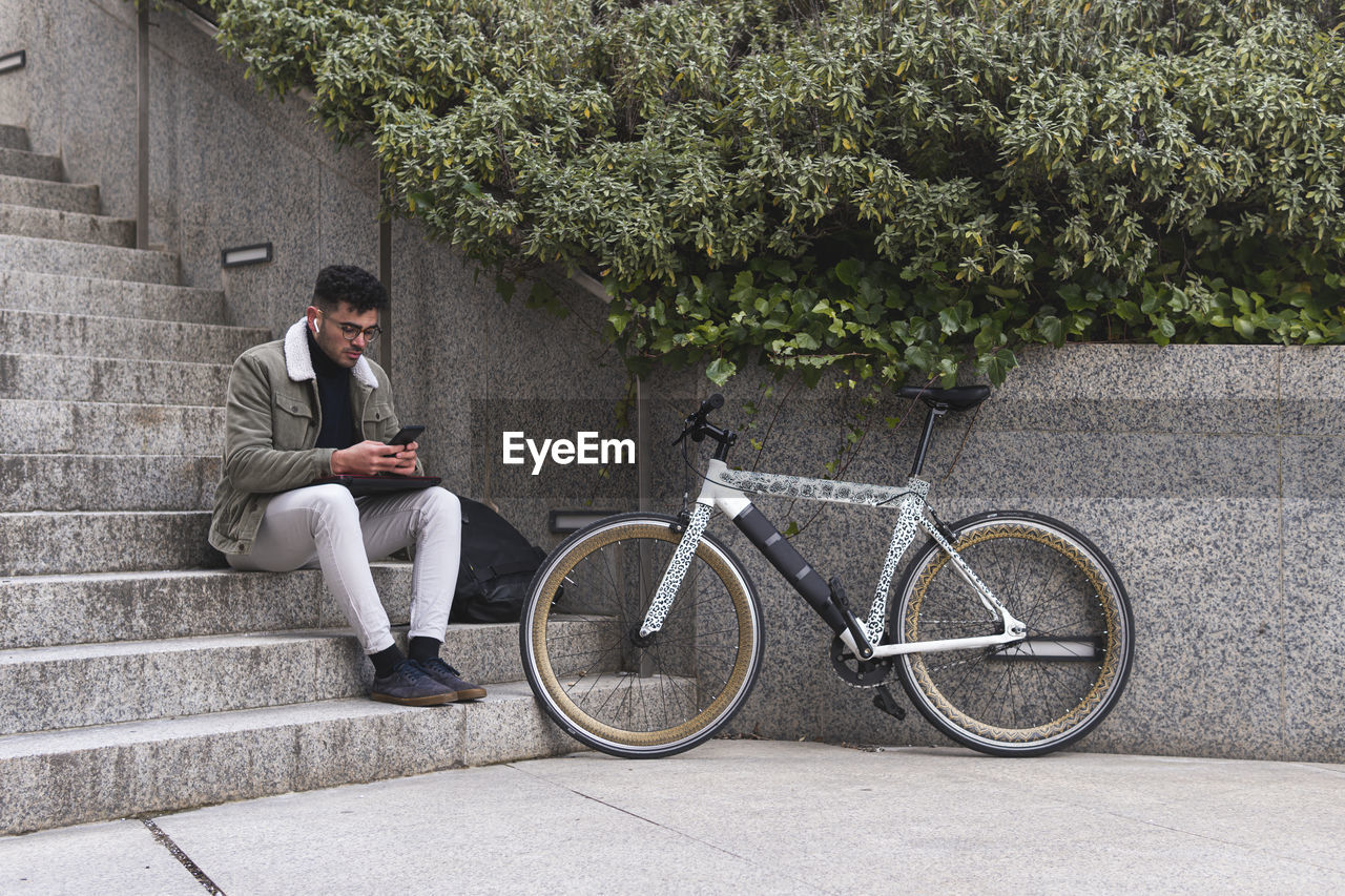 Young man with bicycle using smart phone while sitting on steps