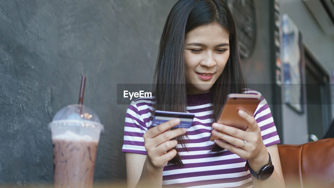 Smiling young woman holding credit card while using smart phone at table with coffee in cafe