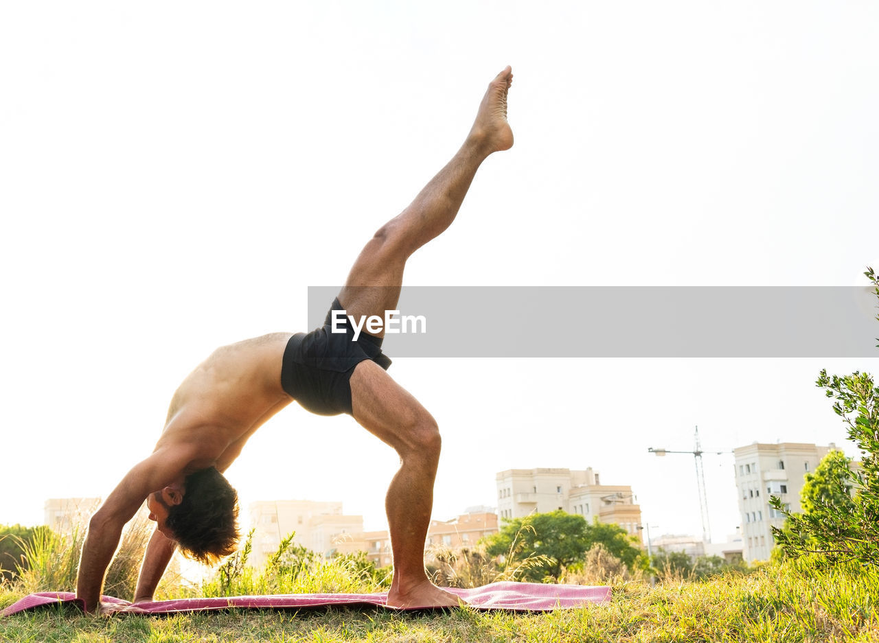 Side view of flexible barefoot man in shorts doing eka pada urdhva dhanurasana pose on mat during yoga session in sunny summer morning in park