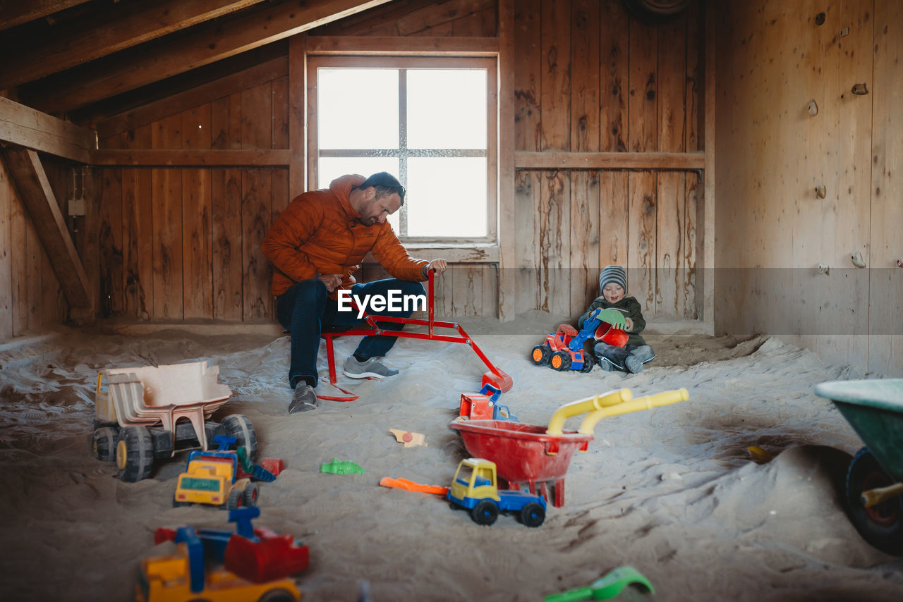 Father and son playing in roofed outdoor sandbox with digger and truck