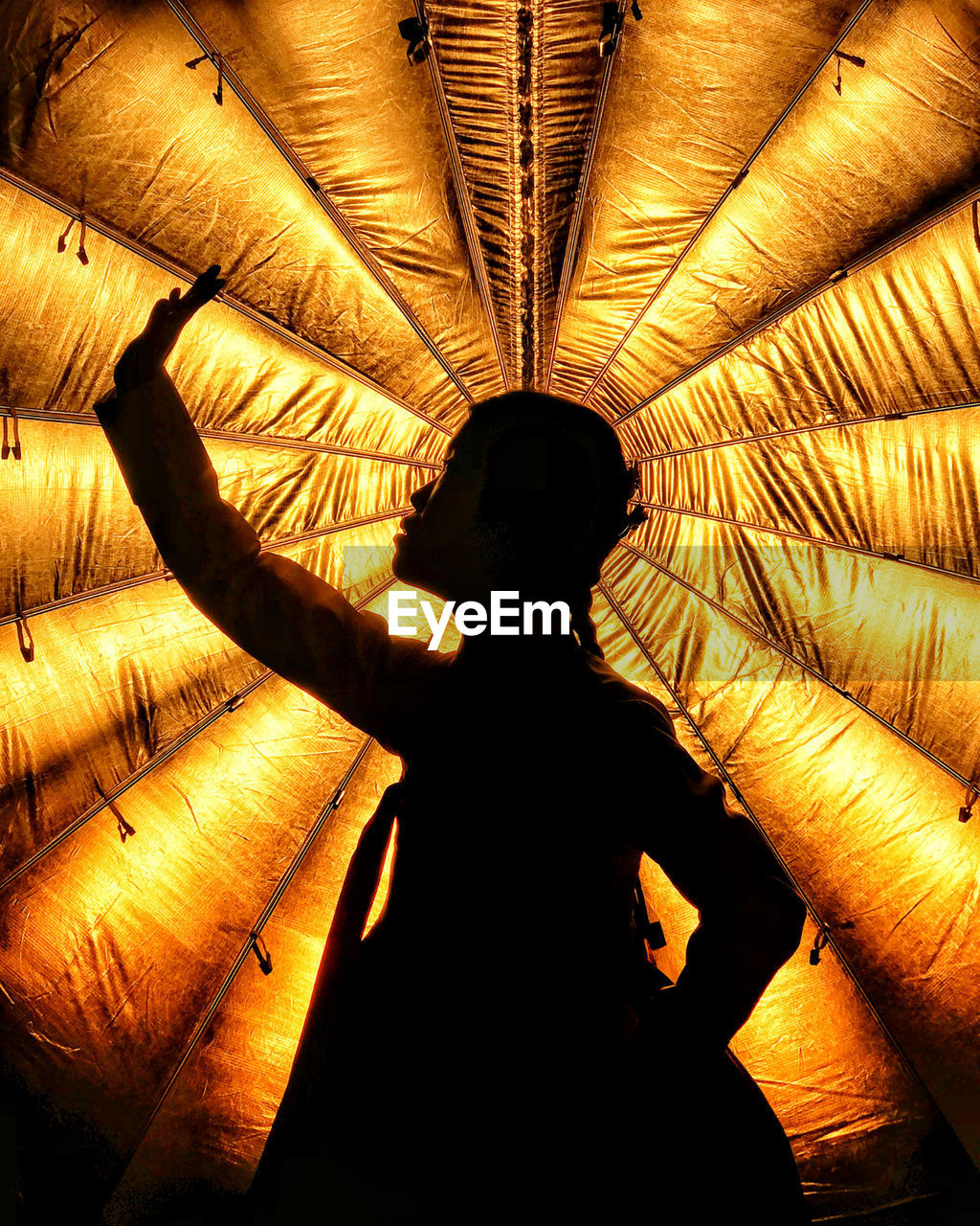 Silhouette woman with hand raised standing against illuminated orange curtain