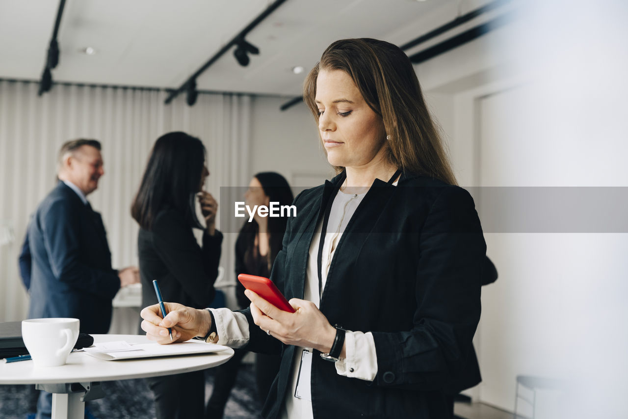 Female business person using phone and writing while standing at table in office workplace