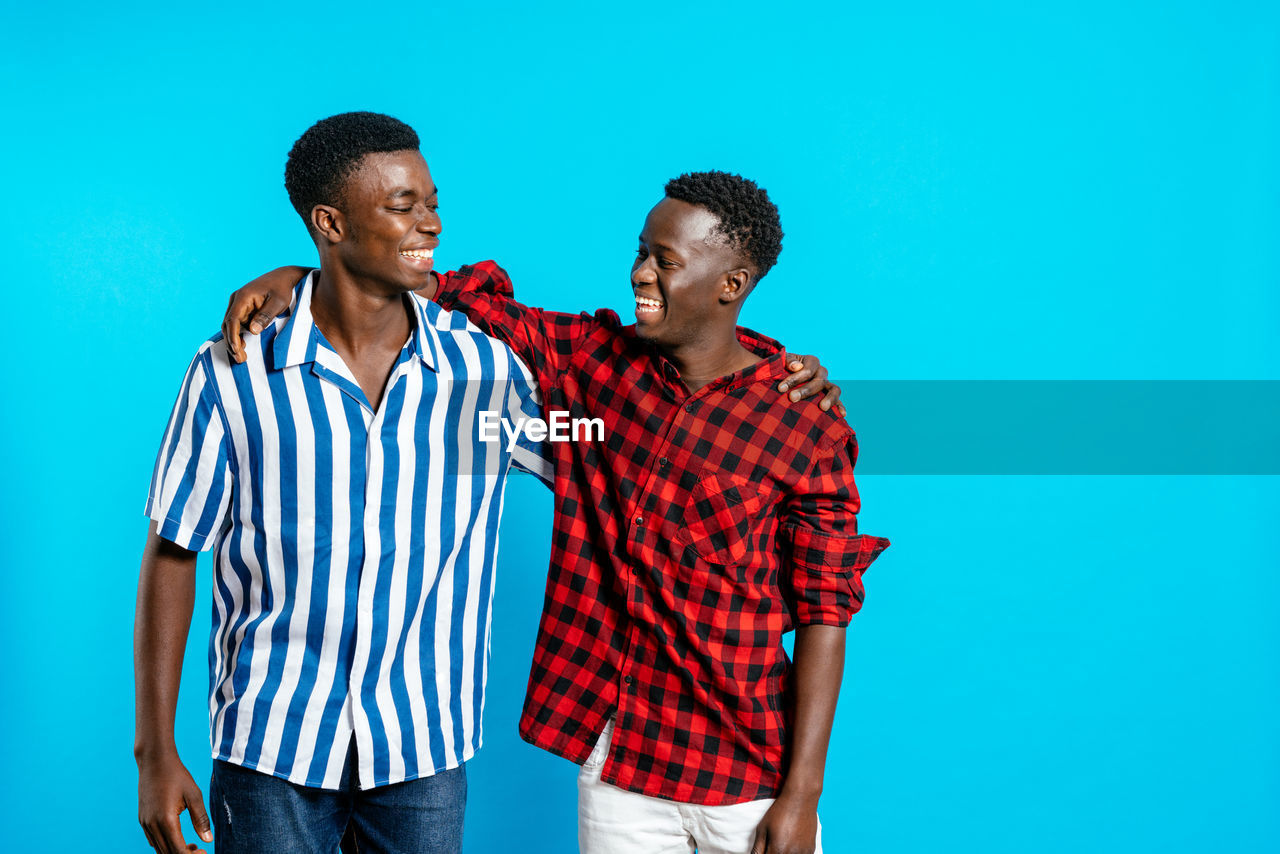 Cheerful african american male friends in stylish outfit standing together on blue background in studio and looking at each other