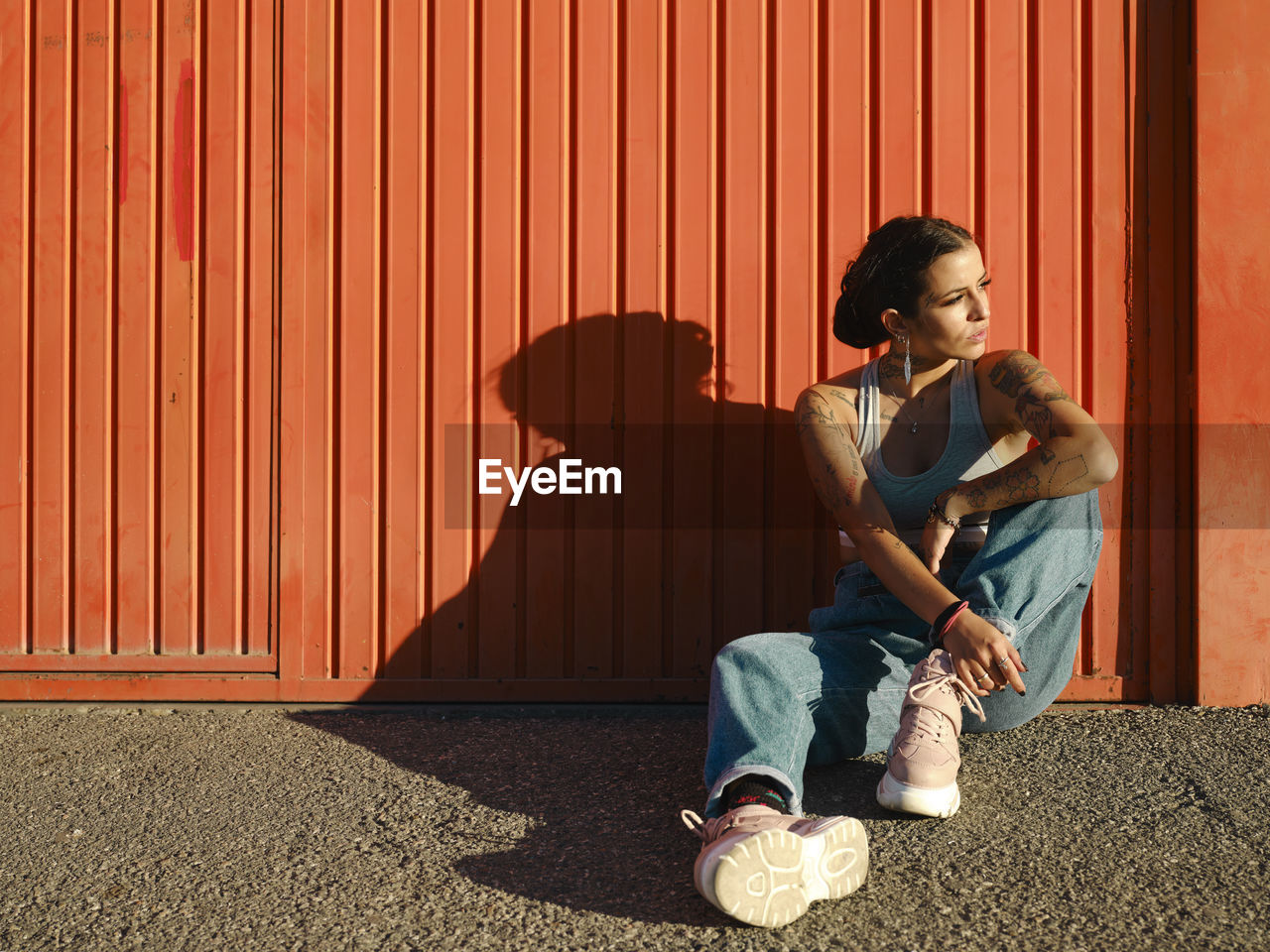 Confident young female with tattoos wearing stylish outfit sitting on floor near red wall and looking away