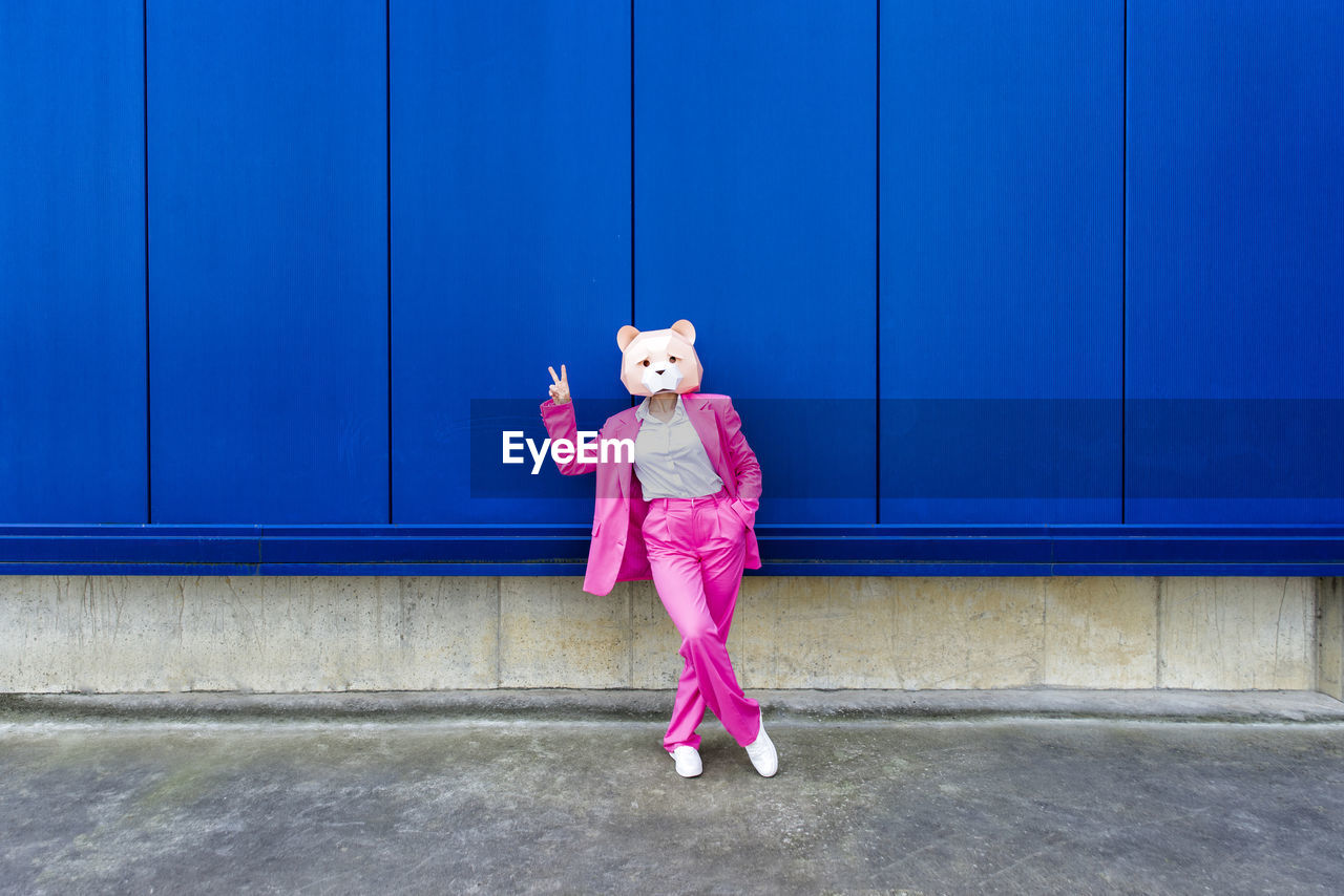 Woman wearing vibrant pink suit and bear mask making peace sign in front of blue wall