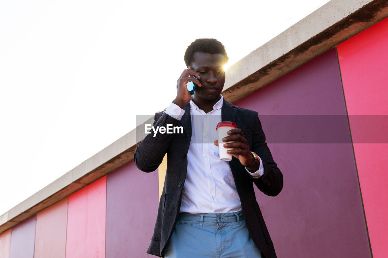 Low angle of serious young african american businessman in trendy clothes holding disposable cup of coffee and discussing business news via smartphone while standing near colorful wall on street