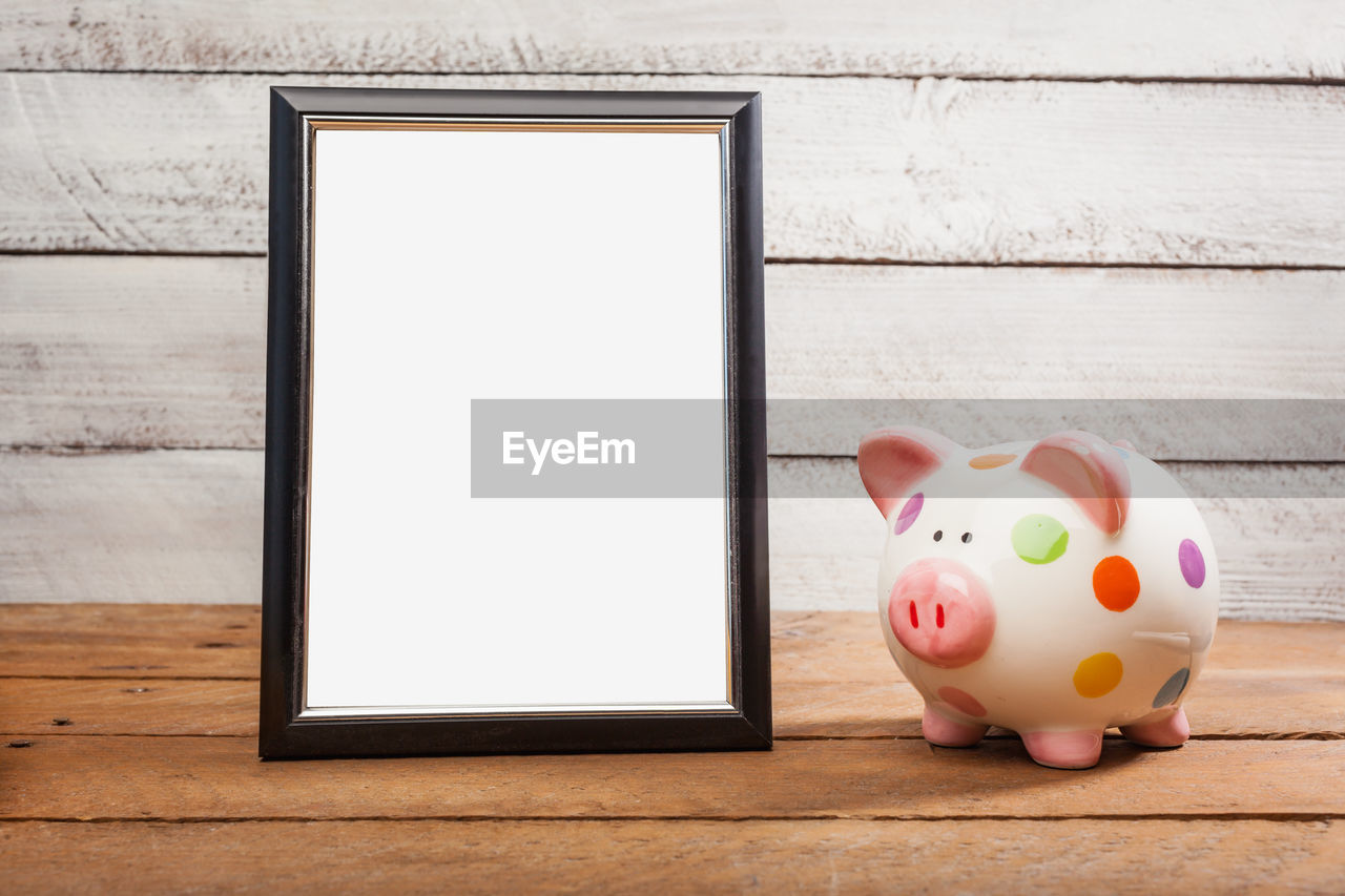 Close-up of blank picture frame with piggy bank on wooden table against wall