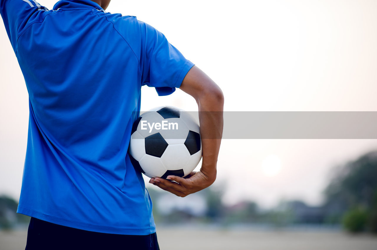 Midsection of man holding soccer ball standing on soccer field against sky