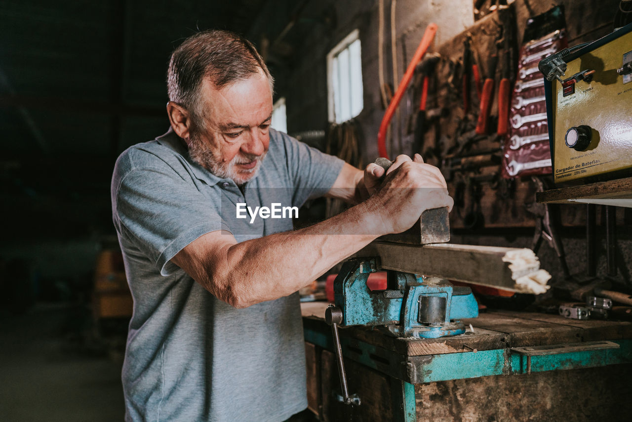 Focused mature male woodworker using jointer for planning wooden plank while working at shabby workbench in joinery workshop