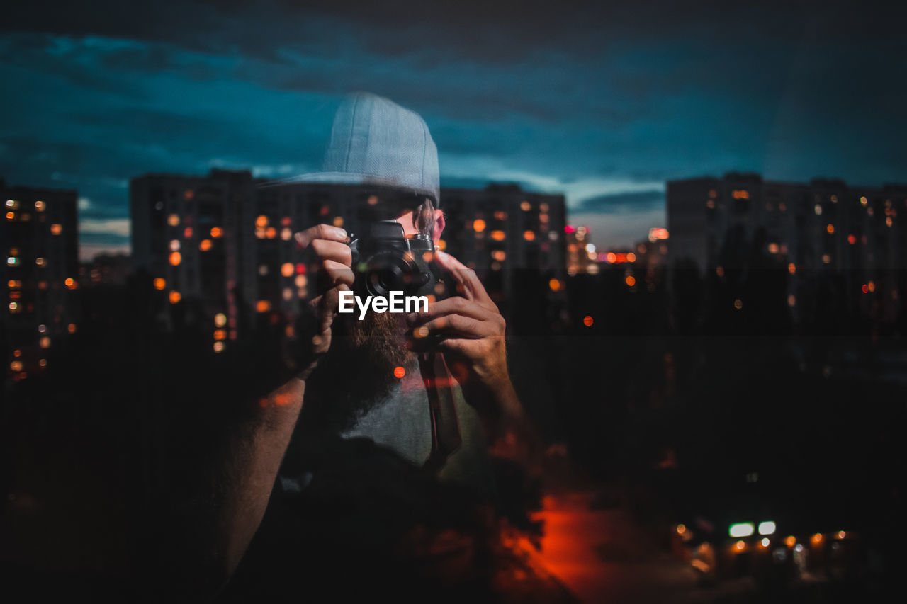 Reflection man photographing illuminated buildings in city on window at night