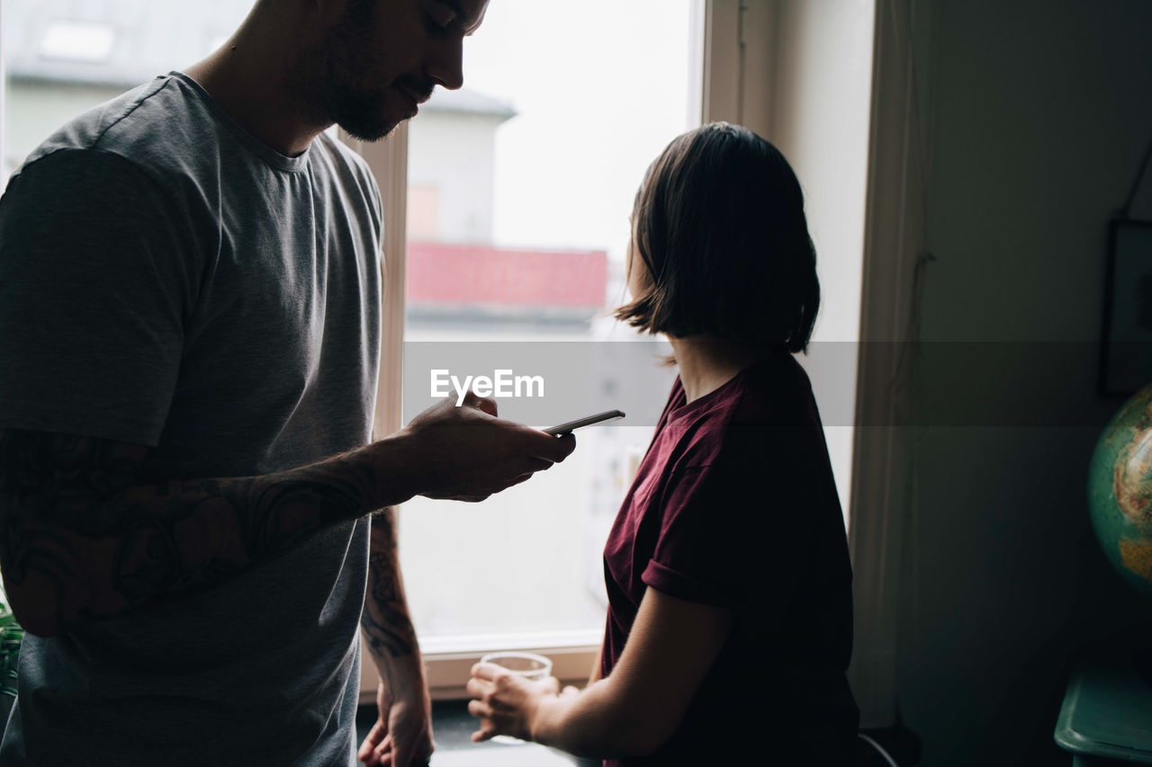 Woman looking through window while man using mobile phone at home