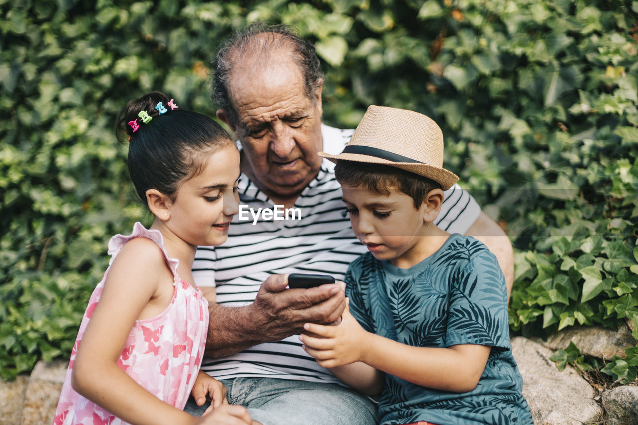 Cute boy wearing hat using smart phone while sitting with grandfather outdoors