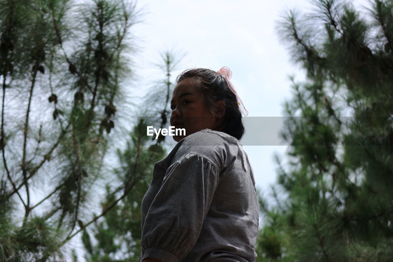 Portrait of young woman looking away against trees