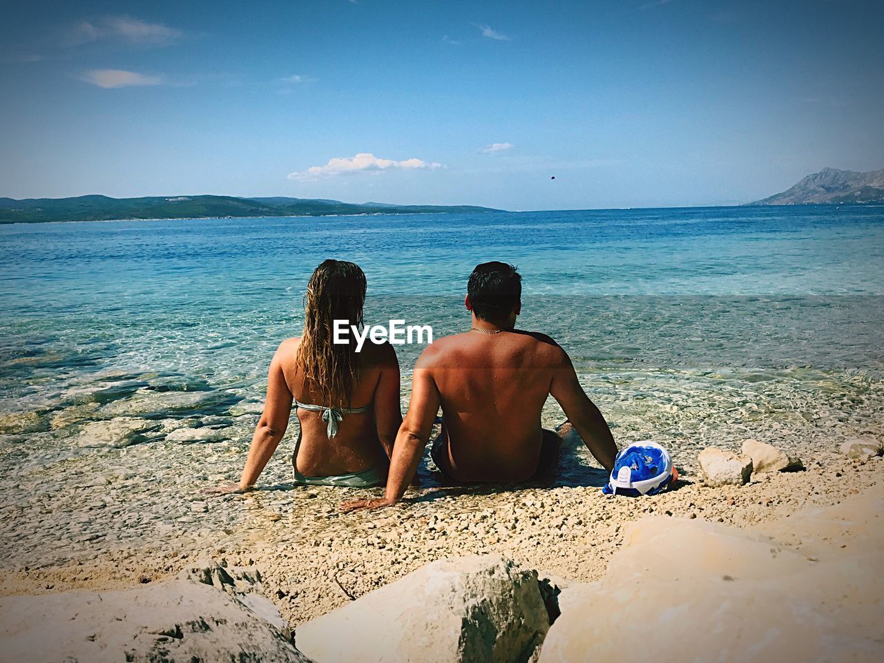 Rear view of couple sitting on shore against blue sky