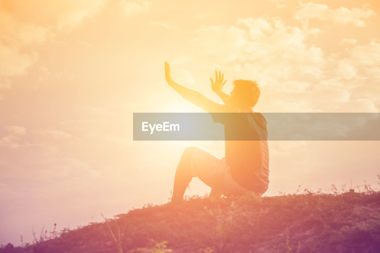 Side view of man gesturing while sitting on land against sky during sunset