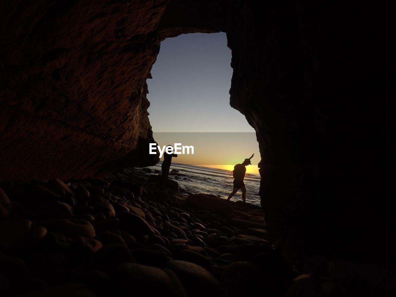 Silhouette couple standing by cave in sea against sky during sunset