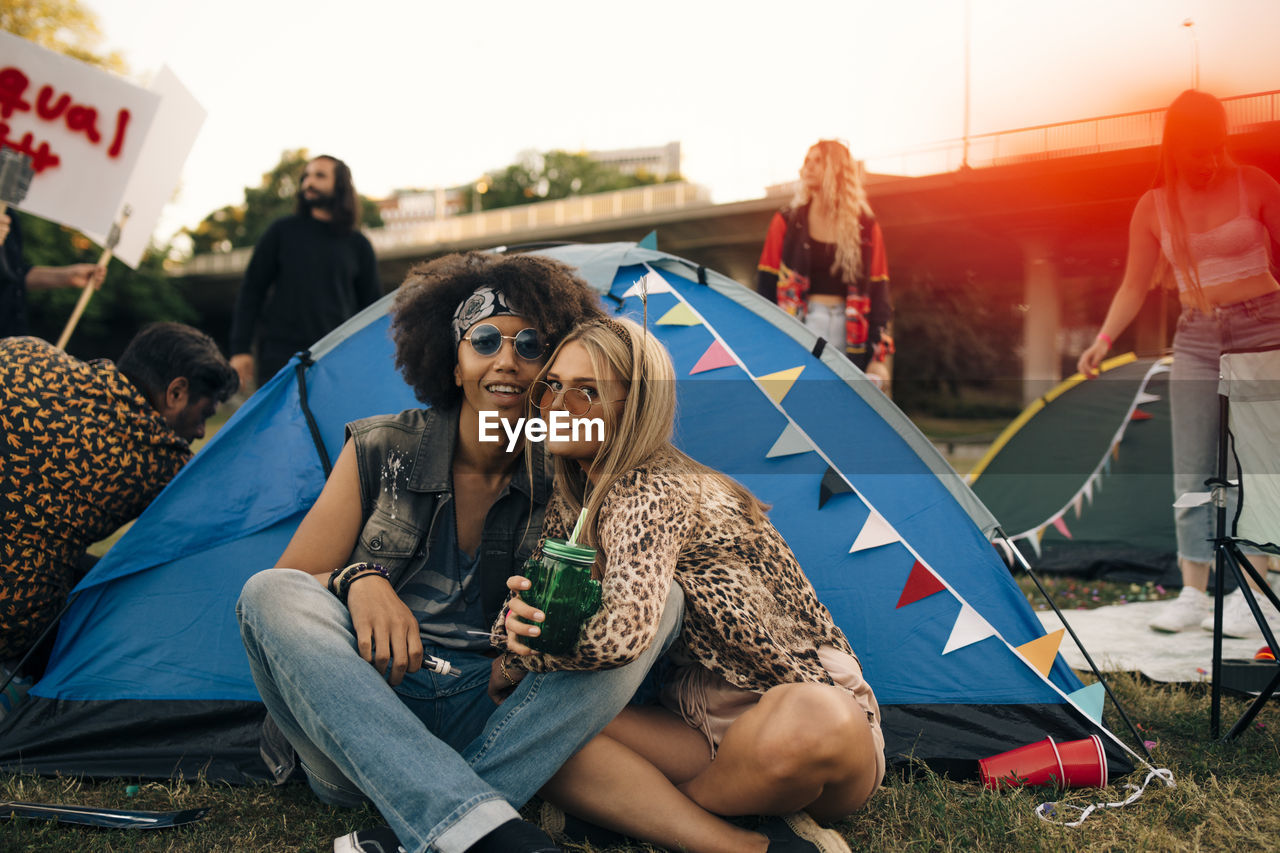 Portrait of smiling friends sitting outside tent on lawn at musical event