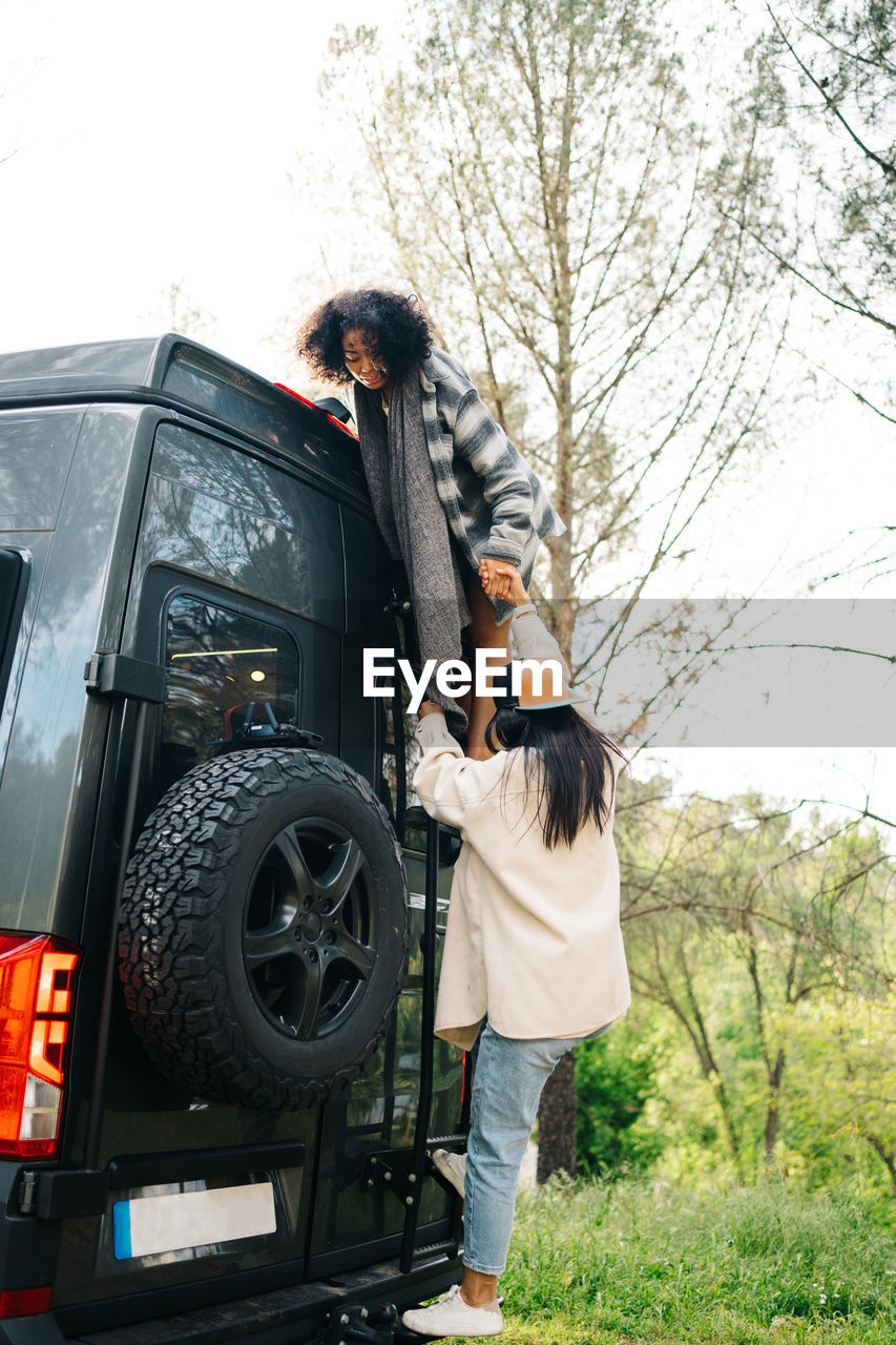 Side view of young african american woman helping friend climbing up camper van parked in green forest during summer adventure in countryside