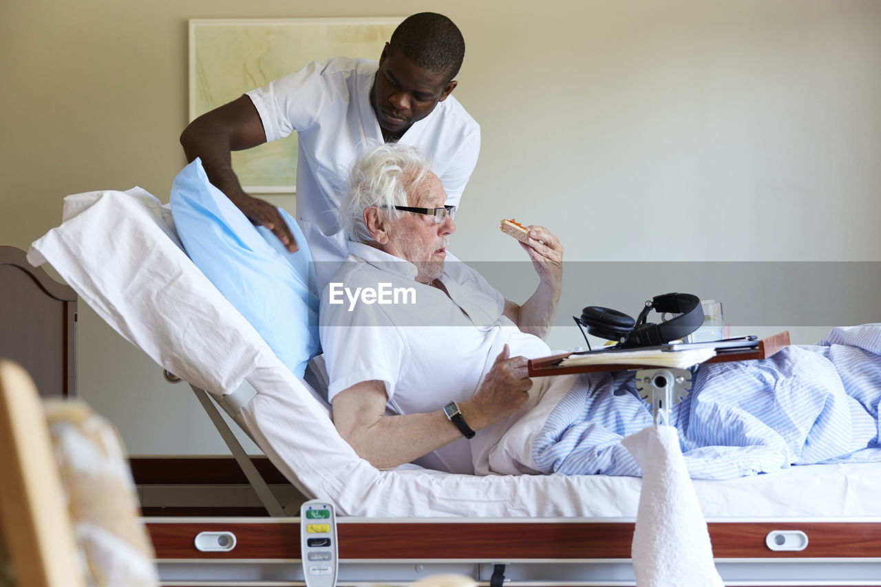 Male nurse adjusting pillow for senior man on bed in hospital ward