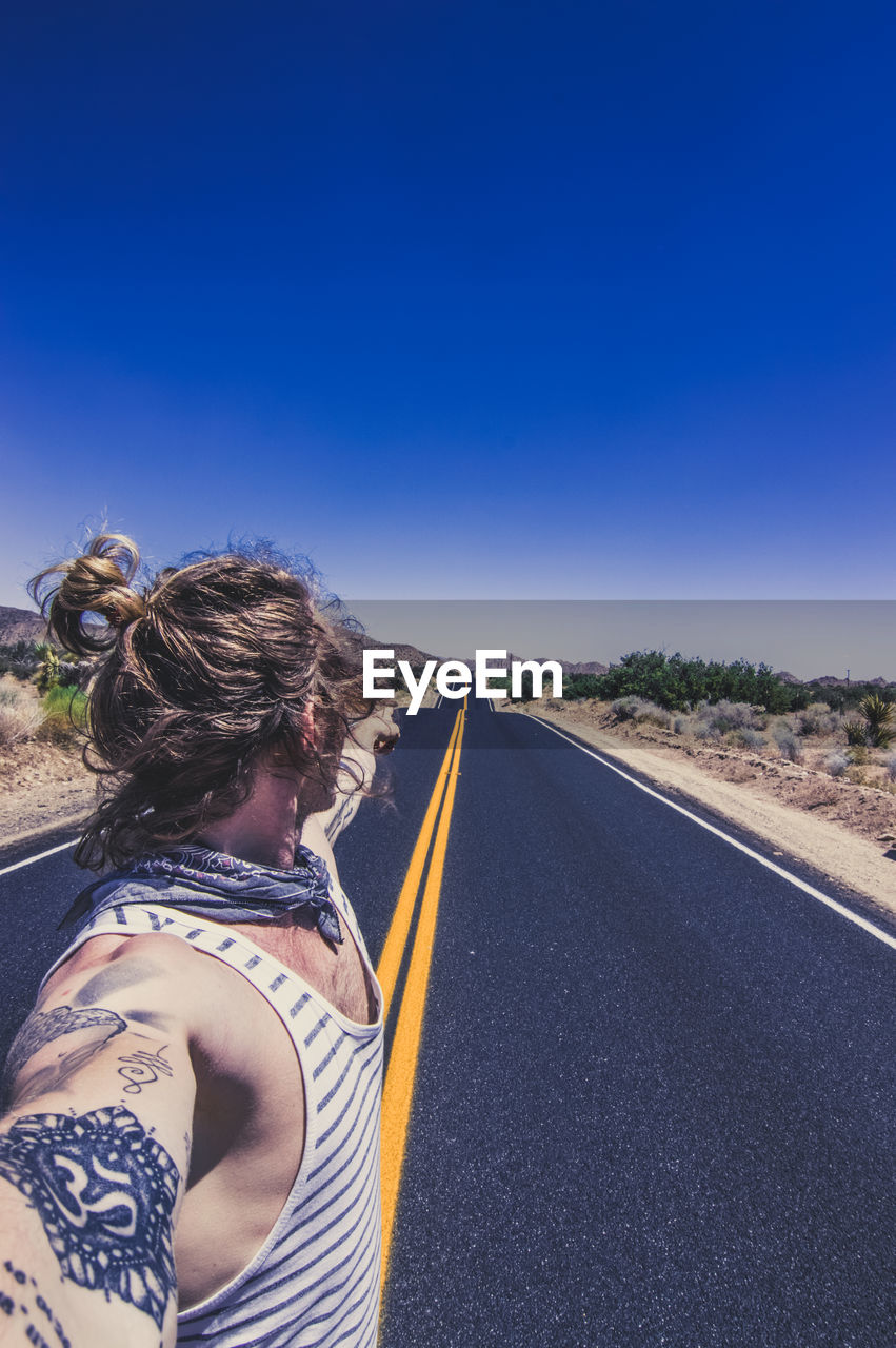 Side view of man pointing at highway against blue sky