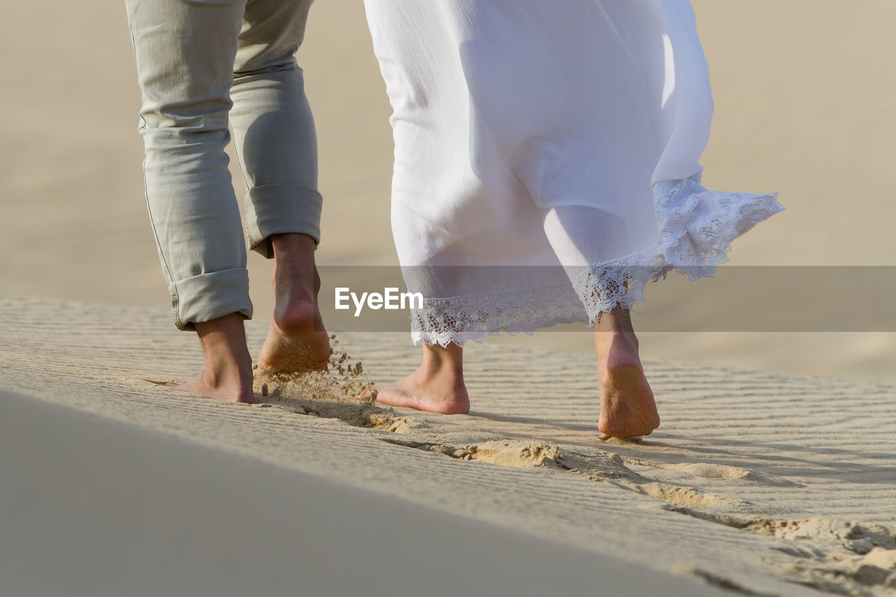 Low section of two  people walking on sand against sky