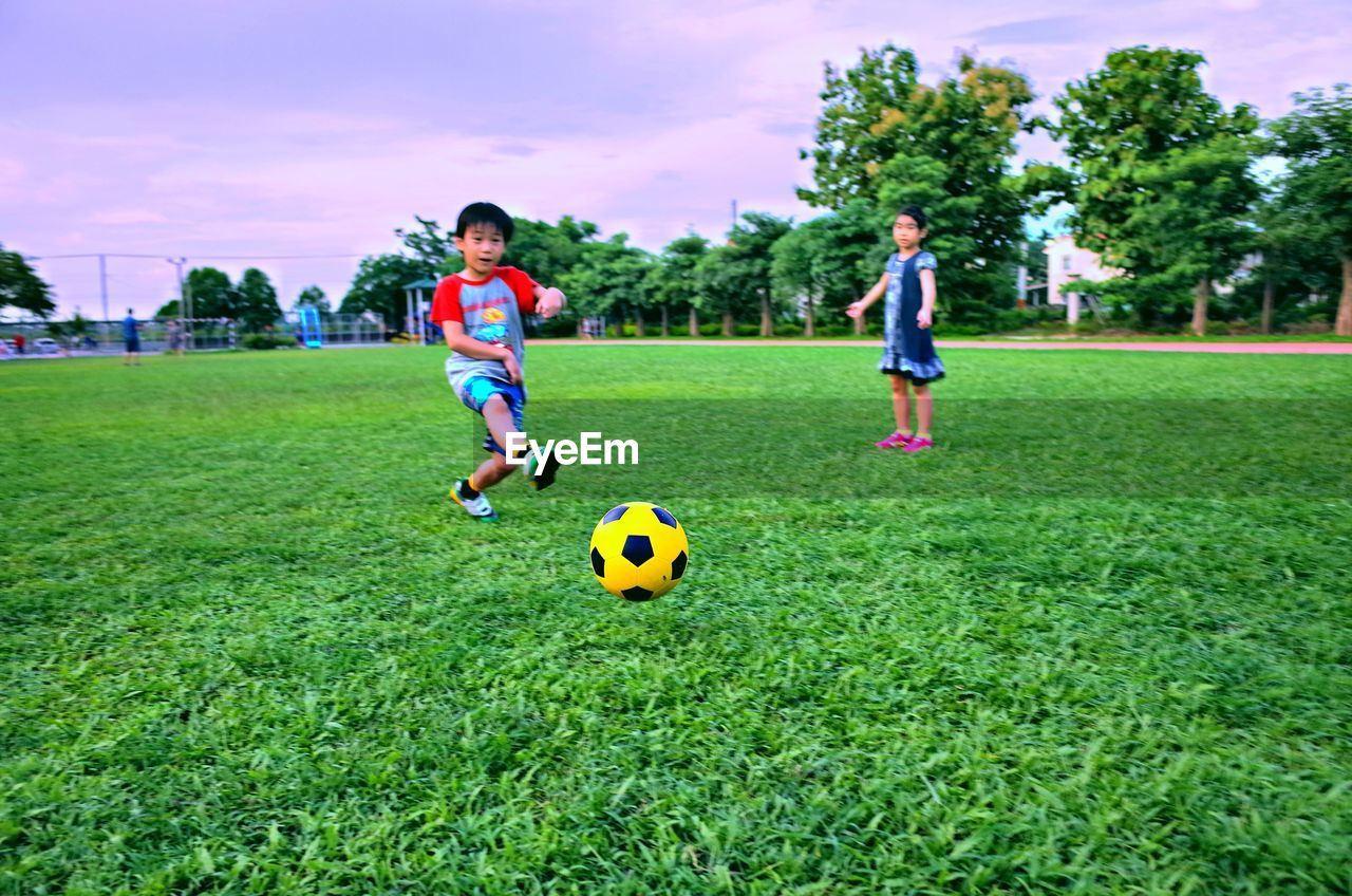 Full length of boy and girl playing soccer with ball on grass against sky