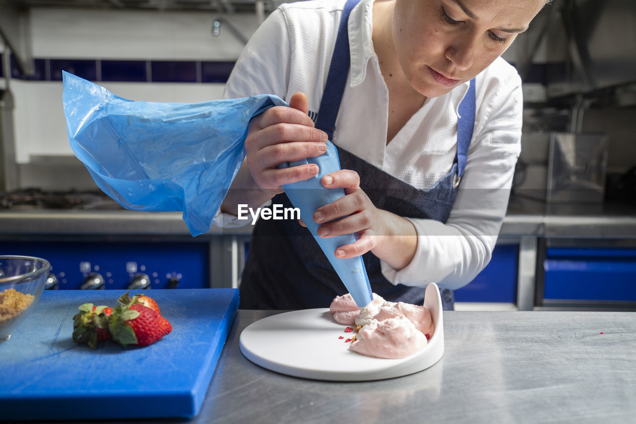 Female chef in apron using pastry bag to squeeze sweet cream on plate while cooking dessert on metal table in restaurant kitchen