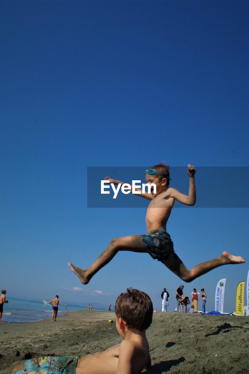 Boys jumping on beach against clear blue sky