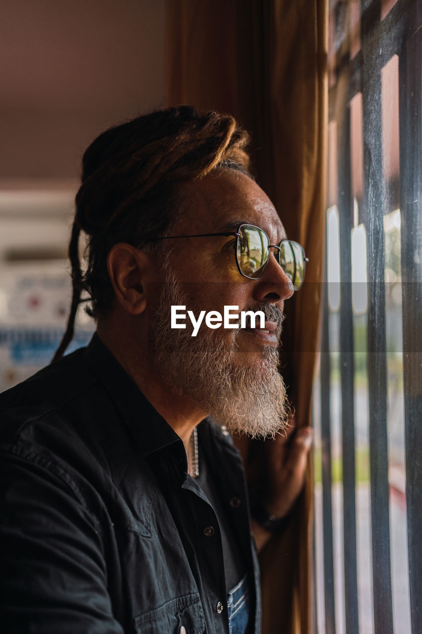 Side view of bearded mature male in casual outfit with sunglasses and dreadlocks standing near window while looking away thoughtfully in light room