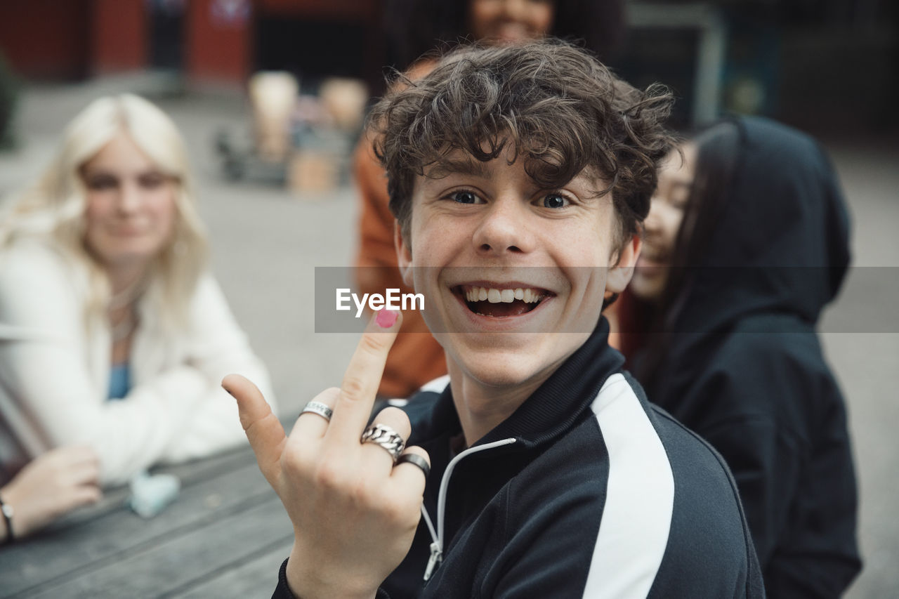 Portrait of happy teenage boy doing obscene gesture while sitting with friends