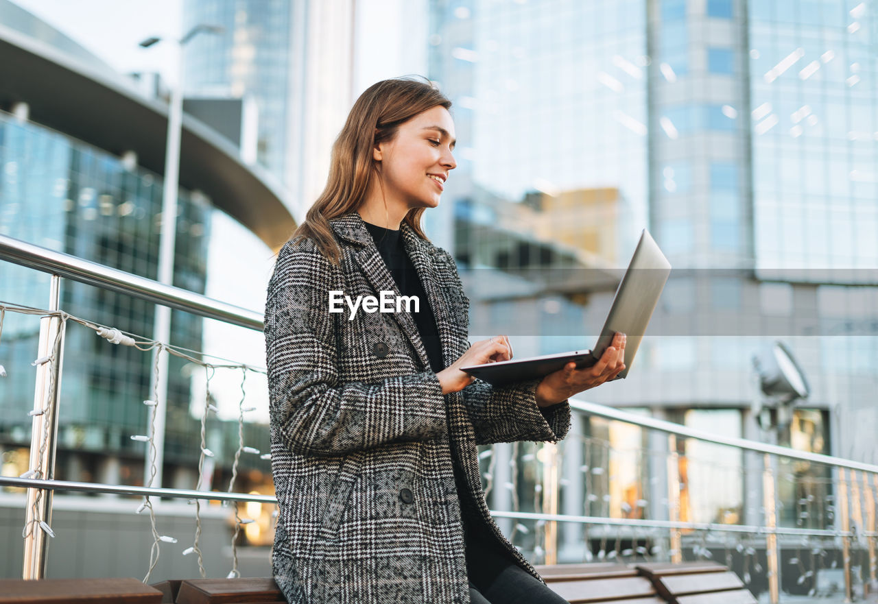 Young smiling woman in coat using laptop in evening city street