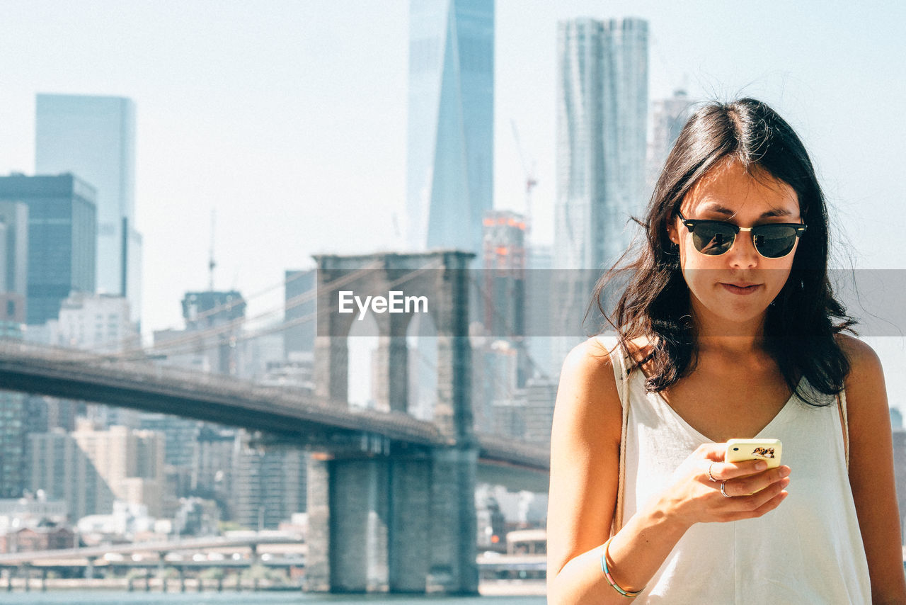 Young woman using smart phone against brooklyn bridge