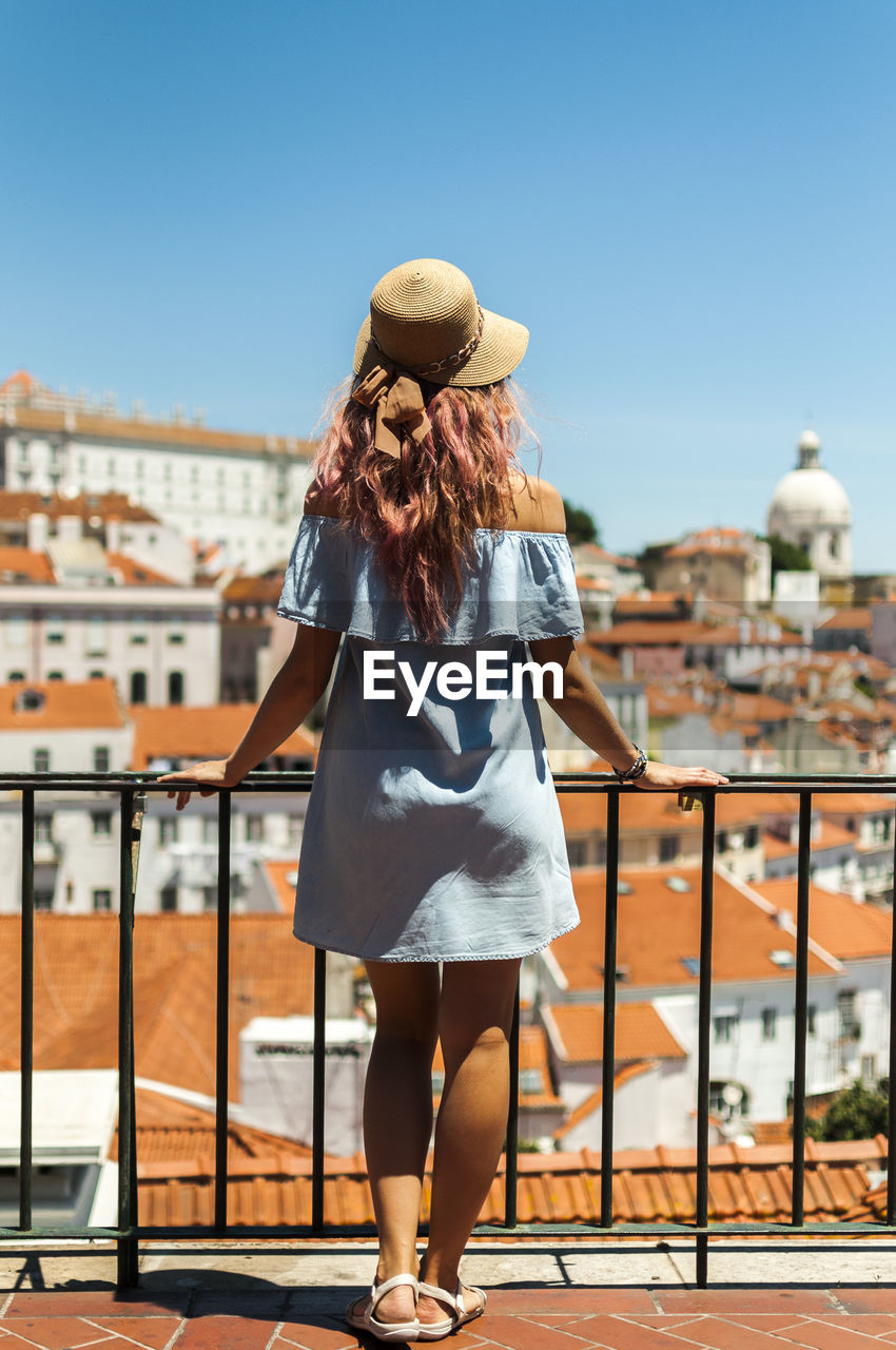 Rear view of young woman looking at buildings while standing by railing in balcony against clear sky