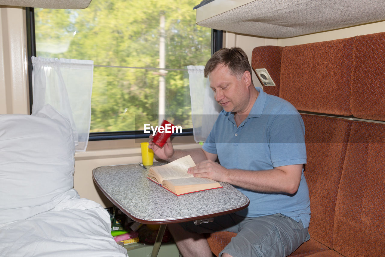 Adult blond man sitting in his train compartment, drinking tea, going on holiday.