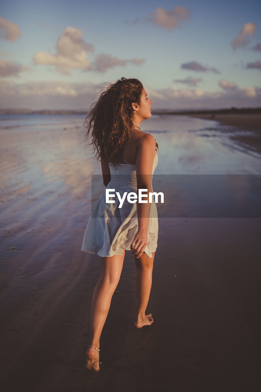 WOMAN STANDING AT BEACH DURING SUNSET