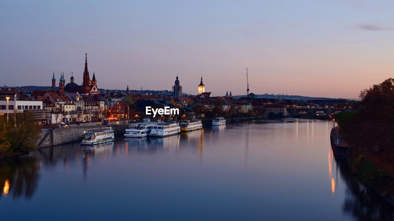 RIVER BY ILLUMINATED BUILDINGS AGAINST SKY AT SUNSET