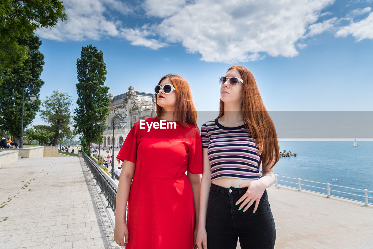 YOUNG WOMAN WEARING SUNGLASSES STANDING ON SHORE AGAINST SKY