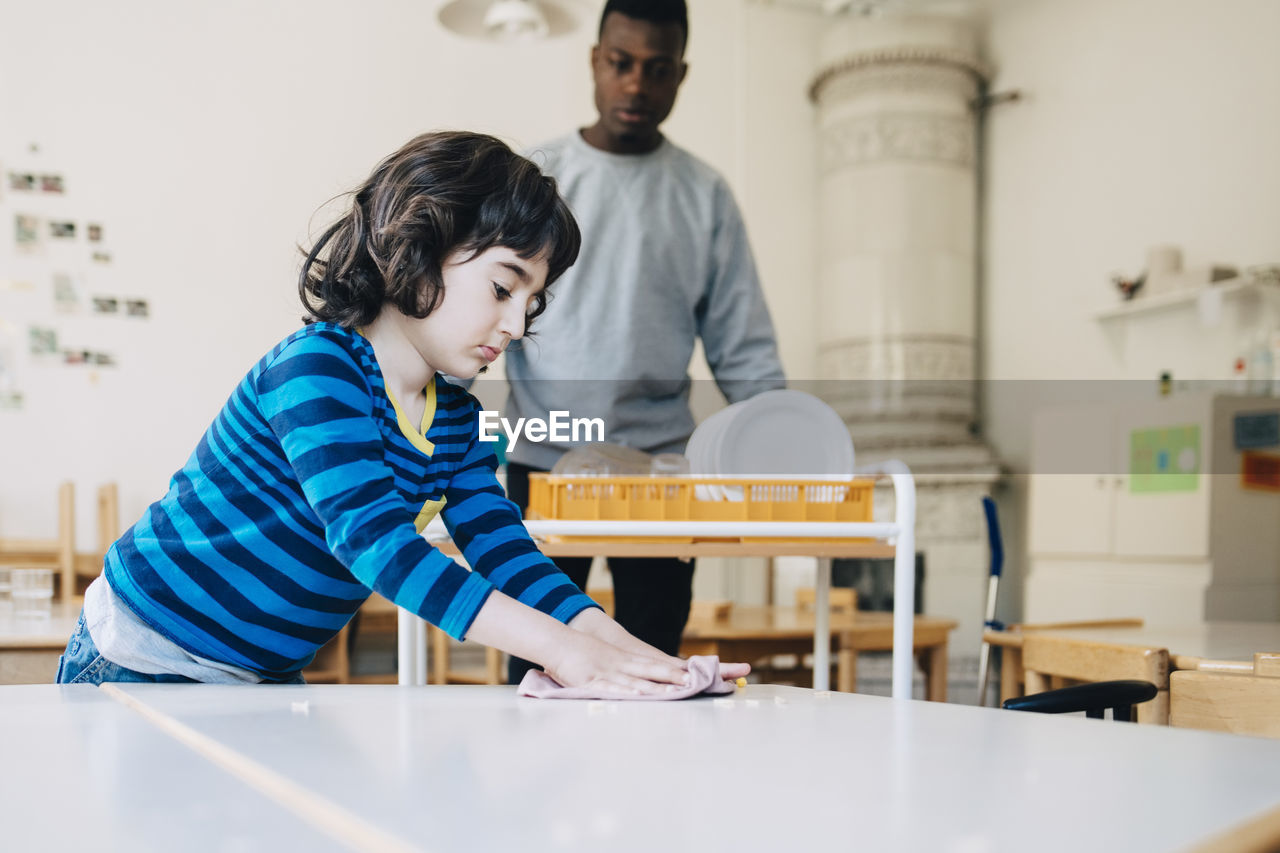 Boy cleaning table with dish cloth while teacher standing in classroom at kindergarten