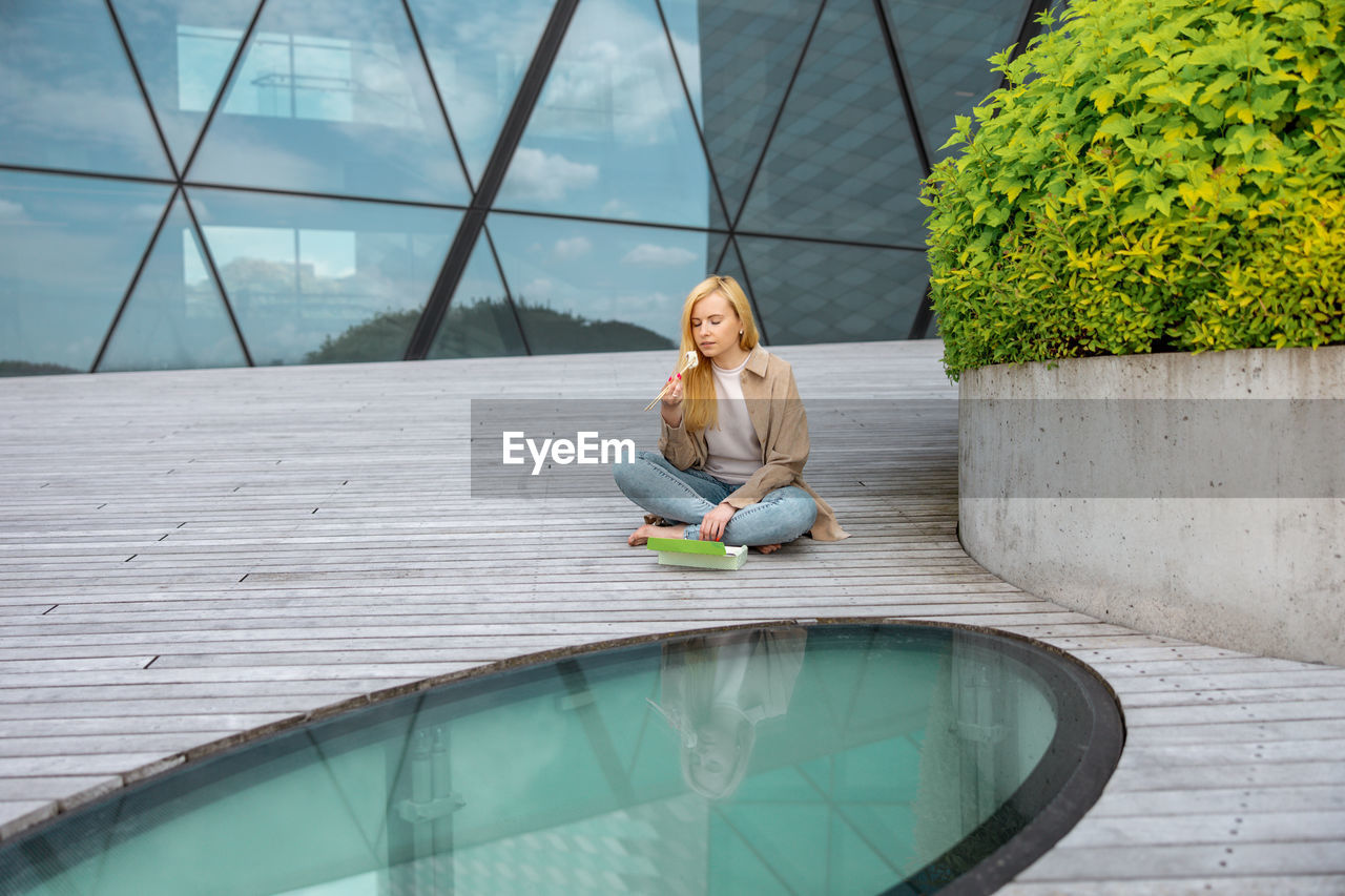 Young beautiful blond woman eating sushi outdoors, on the wooden terrace, by modern building