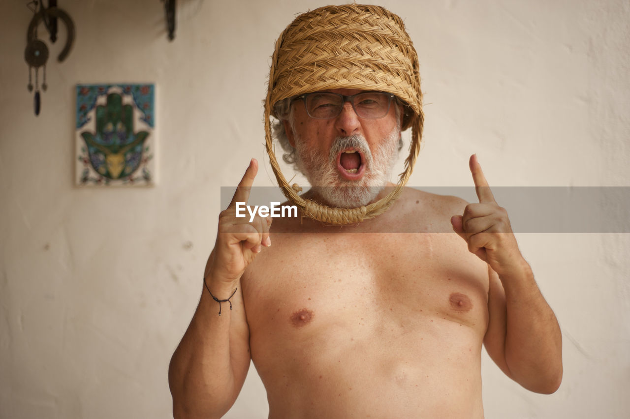 Portrait of shirtless man with basket on head shouting against wall at home