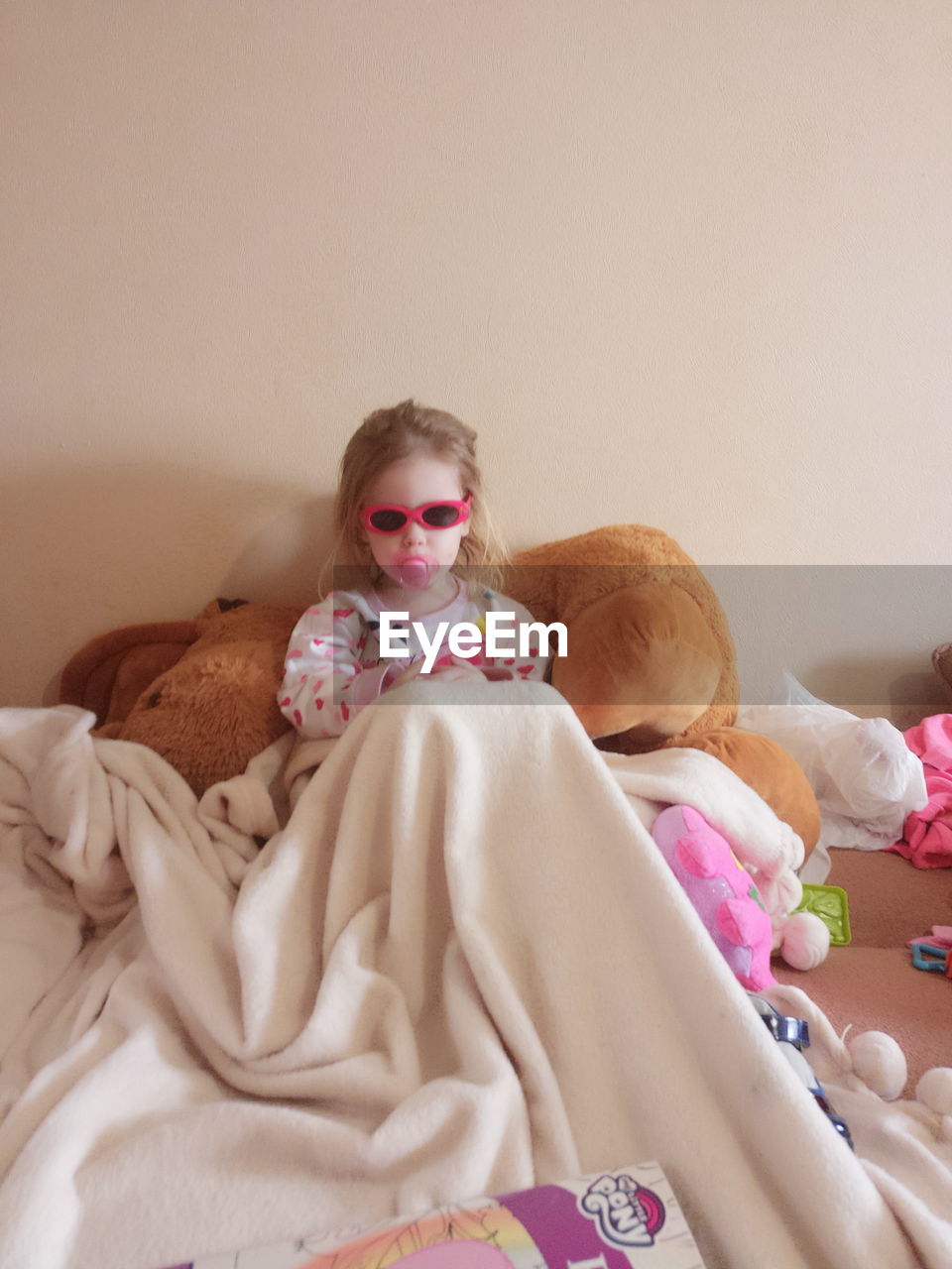 Portrait of girl in sunglasses sucking pacifier while leaning on bed at home