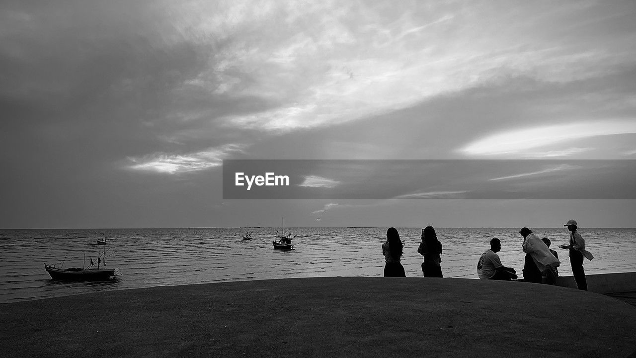 water, sea, sky, horizon, beach, group of people, cloud, black and white, land, monochrome, black, nature, ocean, horizon over water, white, monochrome photography, holiday, beauty in nature, trip, vacation, silhouette, scenics - nature, darkness, men, leisure activity, wave, adult, dusk, coast, women, travel, shore, nautical vessel, outdoors, group, travel destinations, lifestyles, body of water, sand, medium group of people, light, sunlight, evening, tranquility, day, tourism, transportation