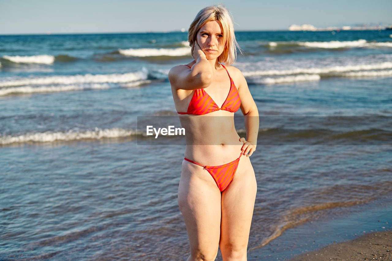 Blond woman in red bikini holding hand on waist and touching neck while standing on beach near waving sea on summer weekend day