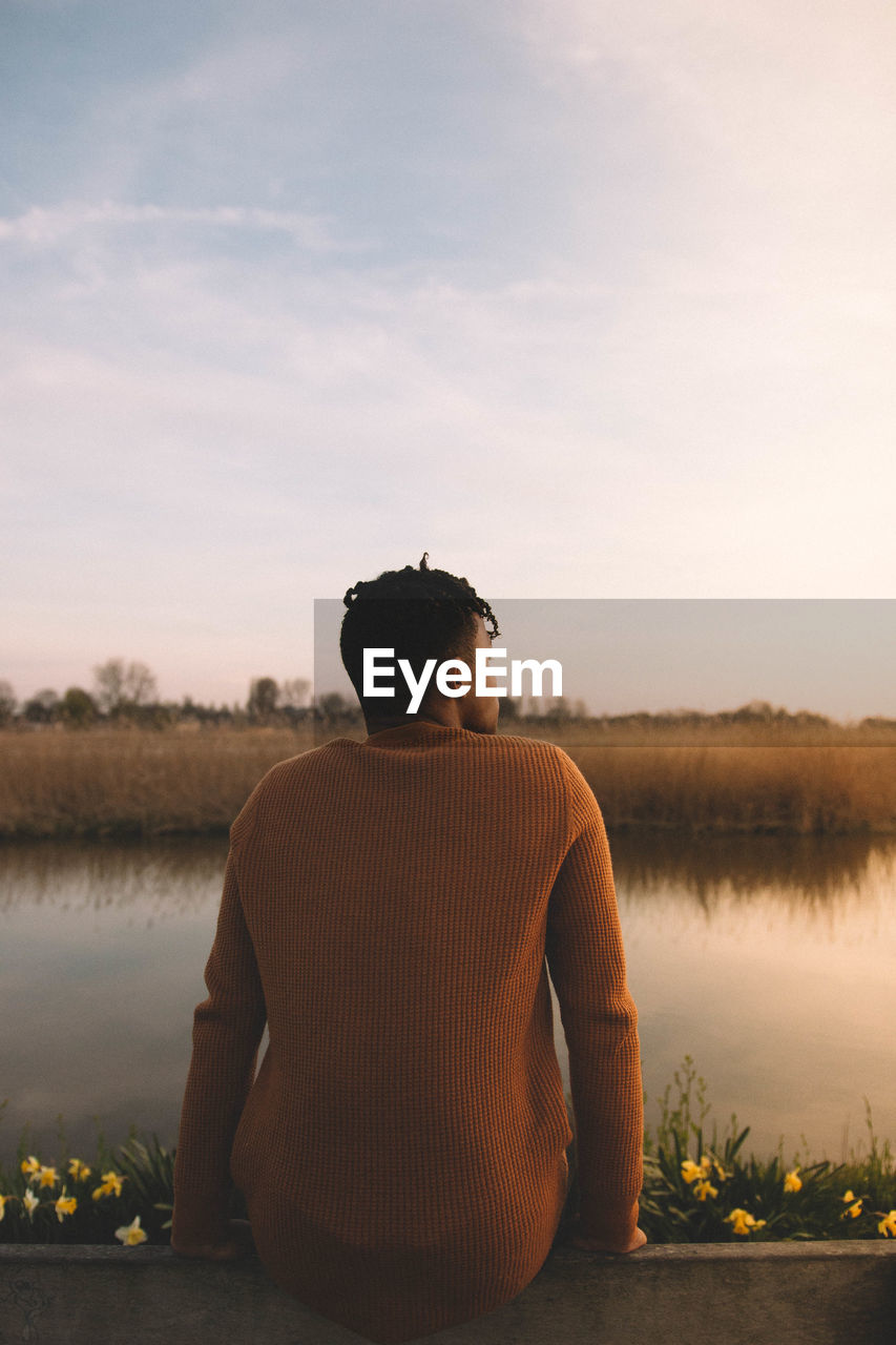 Rear view of man sitting by lake against sky during sunset