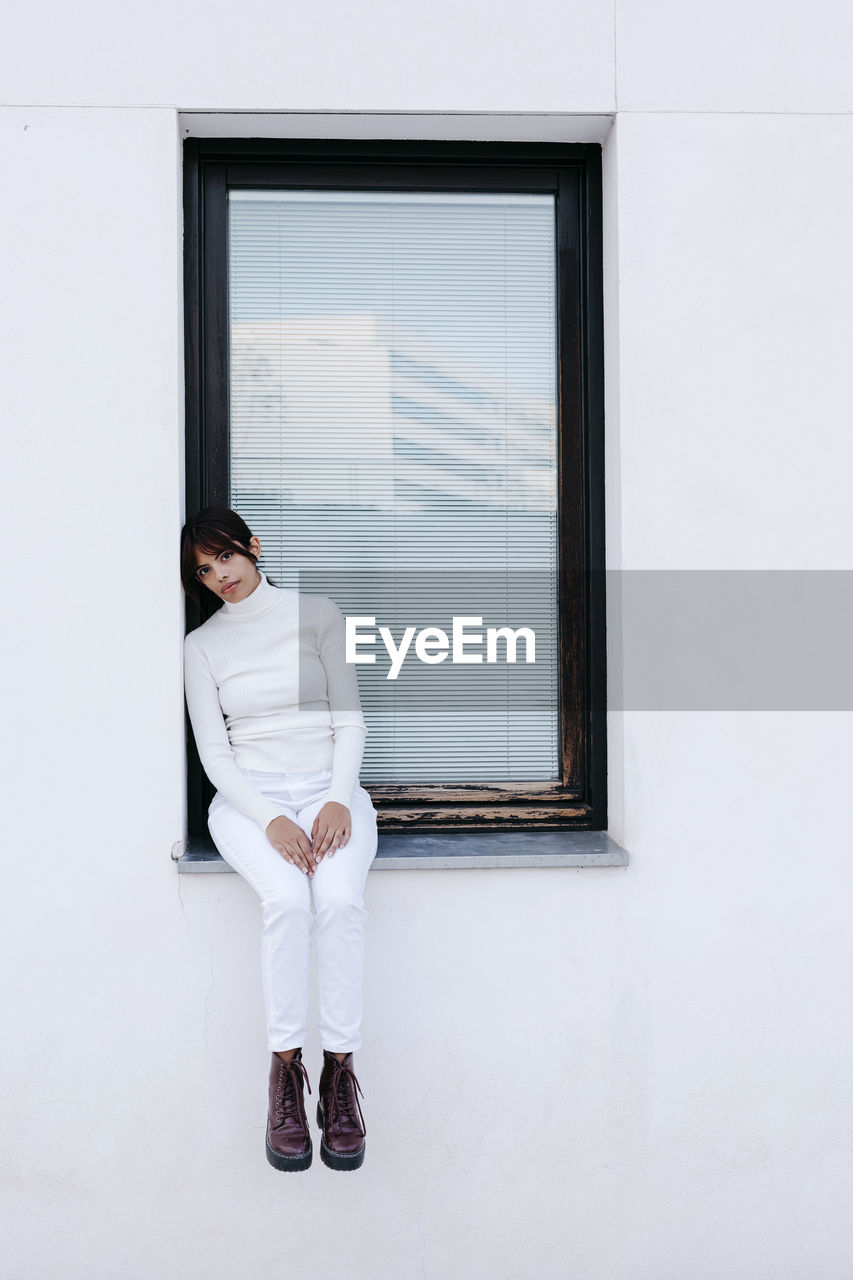 Sad young woman standing on window of white building