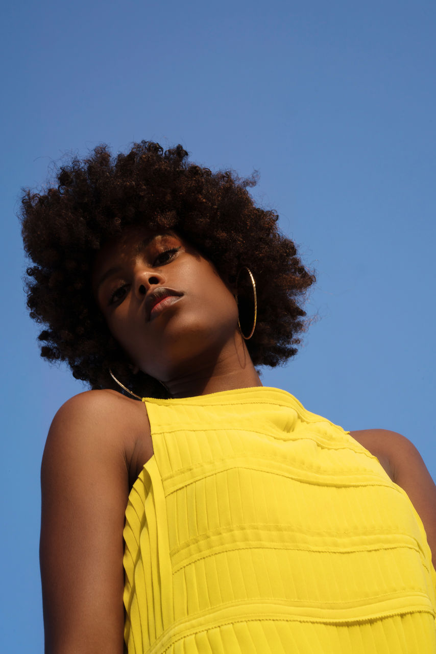 Low angle shot beautiful black woman with afro wearing yellow dress against blue sky