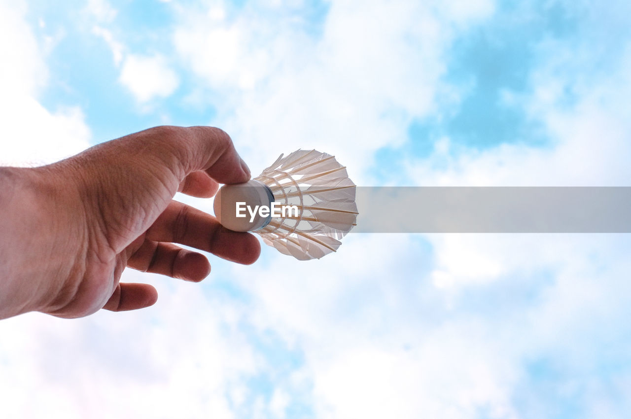 Cropped hand of man holding shuttlecock against cloudy sky