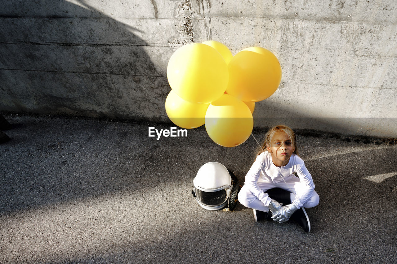 High angle view of girl holding yellow balloon sitting against wall during sunny day