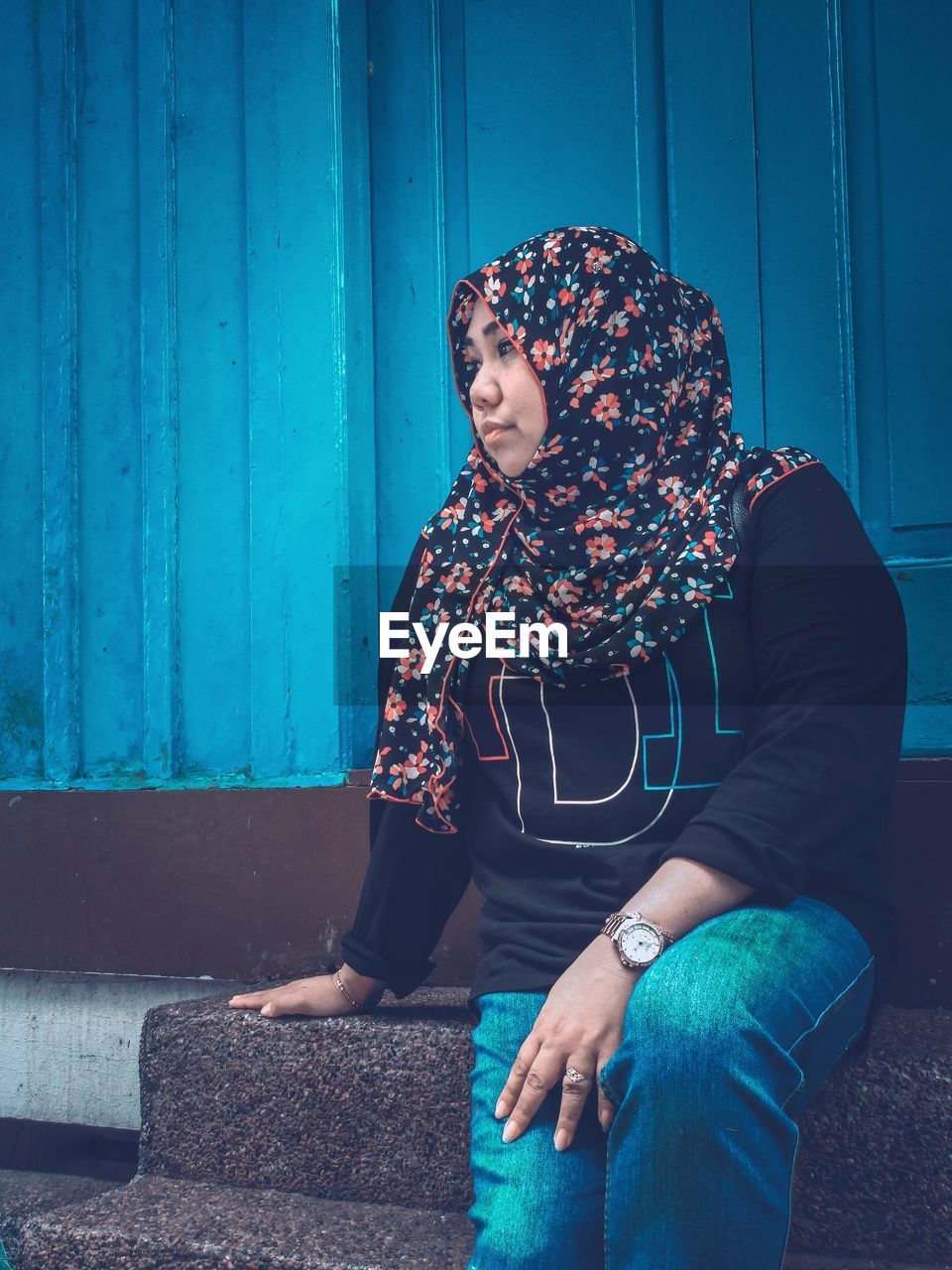 Mid adult woman wearing hijab looking away while sitting on steps against built structure