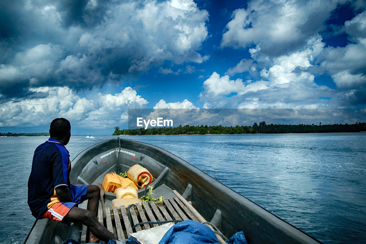 REAR VIEW OF MAN SITTING ON BOAT AGAINST SKY